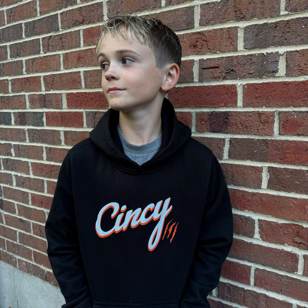 Youth model wearing the black Cincy hoodie in front of a brick wall