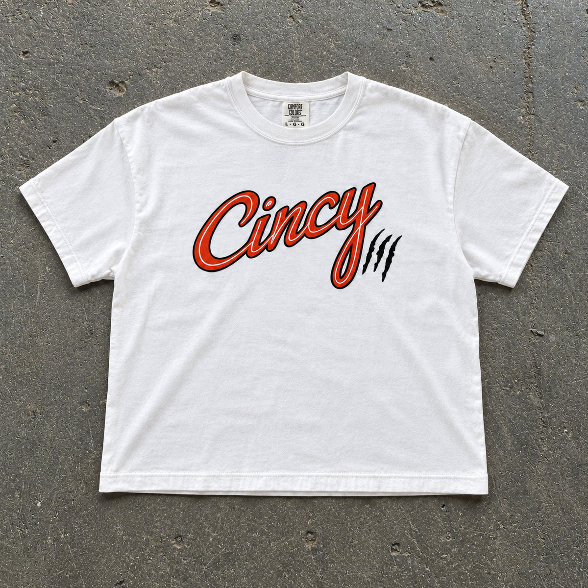 White semi-crop t-shirt with orange Cincy script logo and black claw slashes, flat lay on concrete