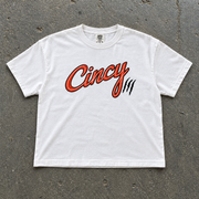 White semi-crop t-shirt with orange Cincy script logo and black claw slashes, flat lay on concrete