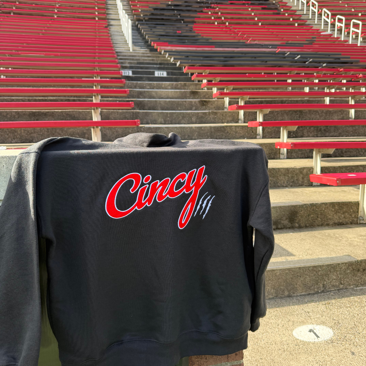 Black mock neck sweatshirt with red Cincy script and white outline, displayed on stadium seating